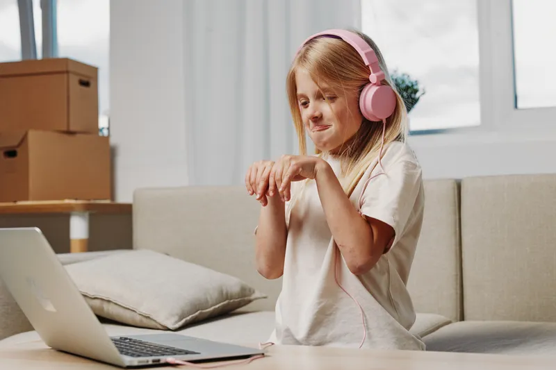 A young girl with long hair wearing pink headphones enjoys music while dancing at home near a laptop on a wooden table in a bright living room. Concept of childhood and music joy.