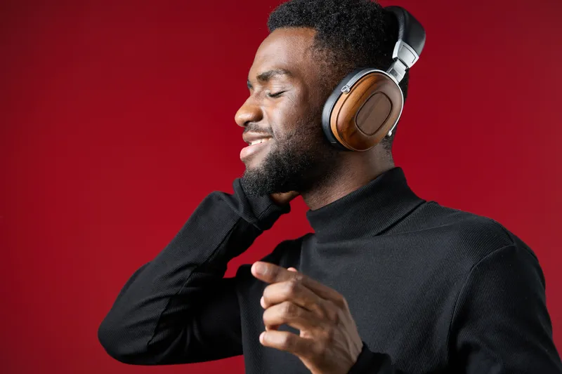 Enjoying music with stylish headphones, a smiling Black man in a black turtleneck against a red background, exuding joy and positivity while listening to beats