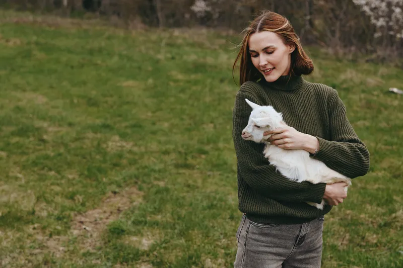 Cheerful woman outdoors with a gentle goat kid, embracing nature and greenery, cozy sweater, sunlit moment, relaxed lifestyle, close bond, joyful interaction, soft breeze, and natural colors