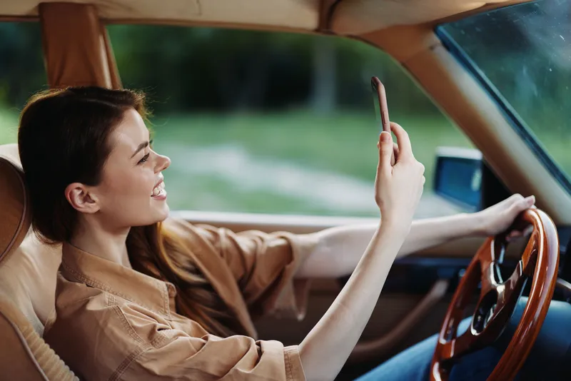 Young woman smiling while taking a selfie in a vintage car, capturing a joyful moment in nature, expressing happiness and leisure during a sunny day.