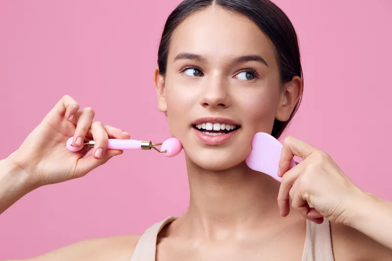 Young woman using facial rollers for skincare, demonstrating a joyful expression against a pink background, promoting beauty and self care routines