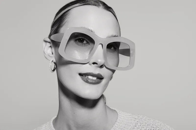 Portrait of a fair skinned Caucasian woman wearing oversized sunglasses and a sweater, smiling confidently against a plain background.
