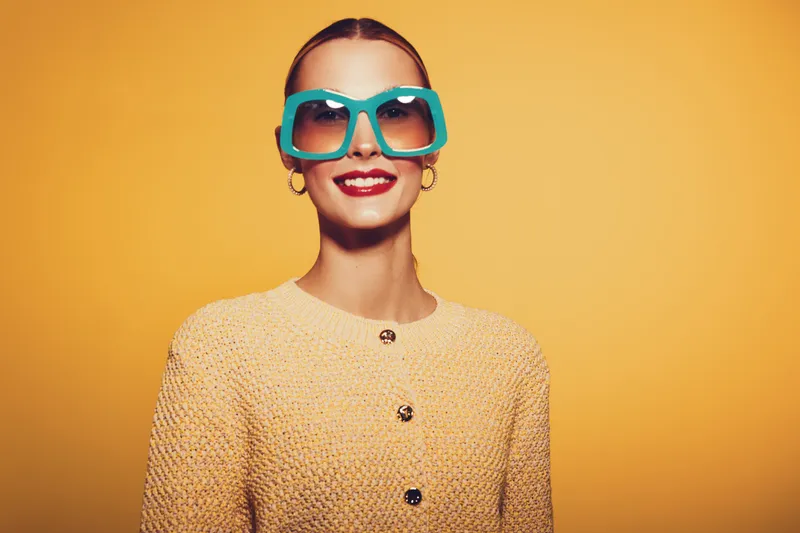 Smiling woman with large blue sunglasses and red lipstick wears textured yellow jacket against bright yellow background. Fashion portrait with retro style and confident mood.