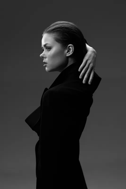 Black and white portrait of a woman with slicked hair wearing a dark coat in profile, showing thoughtful expression and elegant pose.