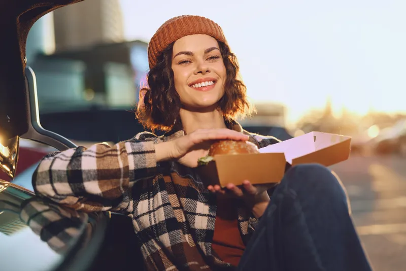 Woman smile with burger by car trunk at golden hour, candid moment showing authenticity, mindful living and emotional storytelling in urban lifestyle with warm glow.