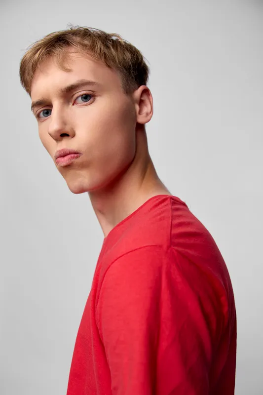 Young man in a red shirt stands in a studio with a slight side glance, showcasing modern fashion mood against a neutral background, ideal for lifestyle and stock projects
