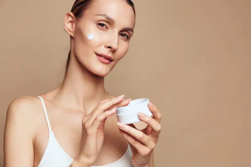 Macro portrait of woman in white top with face cream from cosmetic jar for skincare routine, beauty and hydration, soothing self care, gentle touch on cheek against beige background