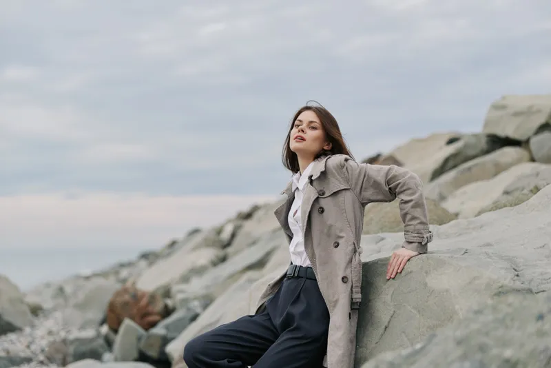 Young woman in patterned coat poses confidently on large rocks by the sea under cloudy sky. Fashion and nature blend to create a calm, stylish outdoor portrait concept.