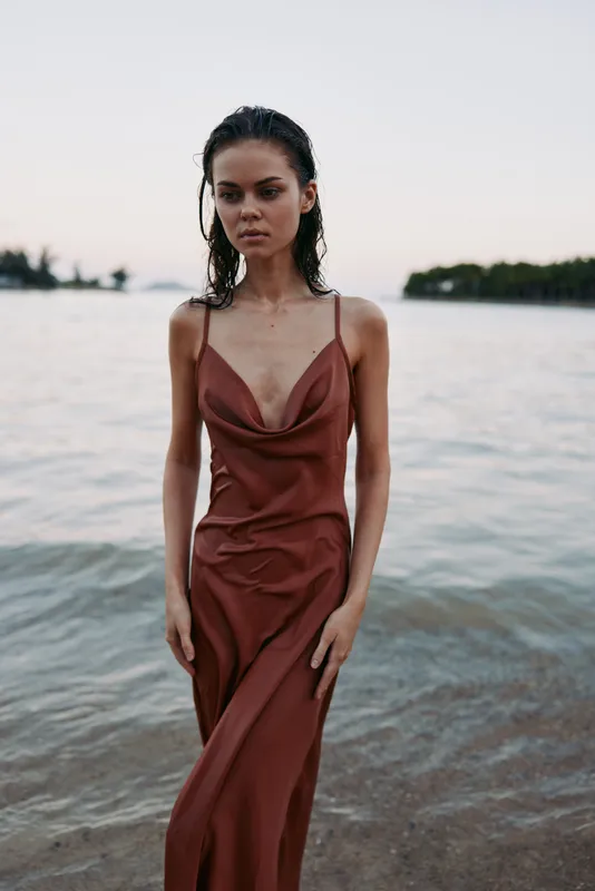 A fashion portrait of a young woman standing at the shoreline in a rust red satin dress, soft light, calm sea, minimal backdrop, and gentle wind across the scene.