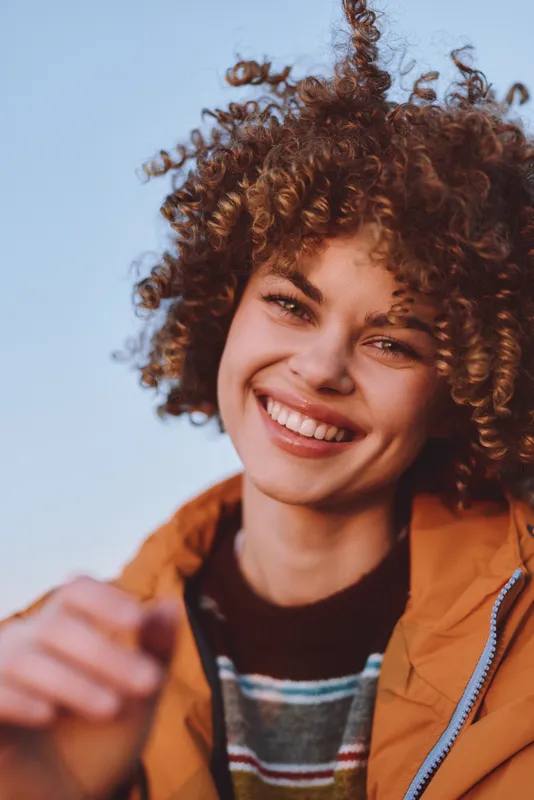 Woman smile curly hair portrait lifestyle outdoors jacket, orange jacket, blue sky background, soft daylight, warm joyful mood, natural look, shallow focus, young female, carefree summer