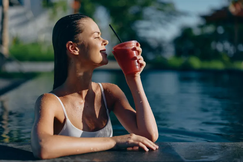 Cheerful woman poolside, sipping a red smoothie through a straw, sun on her skin, relaxed posture, water sparkle, vibrant hues, outdoor lifestyle moment