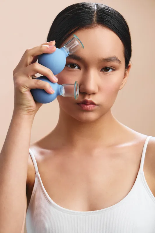 asian woman with dark hair wearing a white tank top holds a blue cupping device to her temple, posing in a clean studio to convey beauty and wellness.
