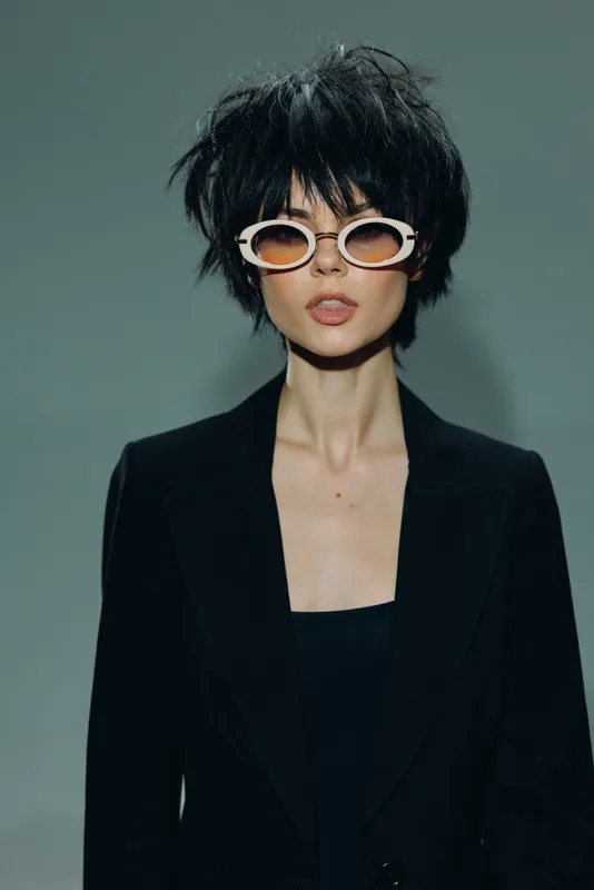 Stylish young woman with short black hair wearing white round sunglasses and a black blazer against a muted background.