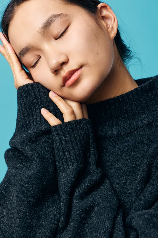 Young woman with closed eyes resting head on hand wearing dark knitted sweater against blue background for relaxation, calmness, emotion and peaceful portrait concept.