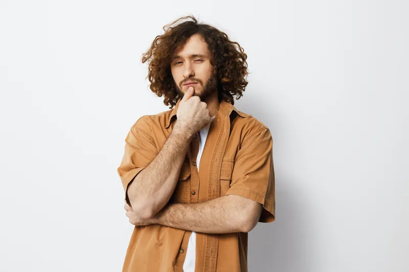 Thoughtful young man with curly hair in a casual outfit, pondering on a light background, expressing contemplation and creativity in his pose