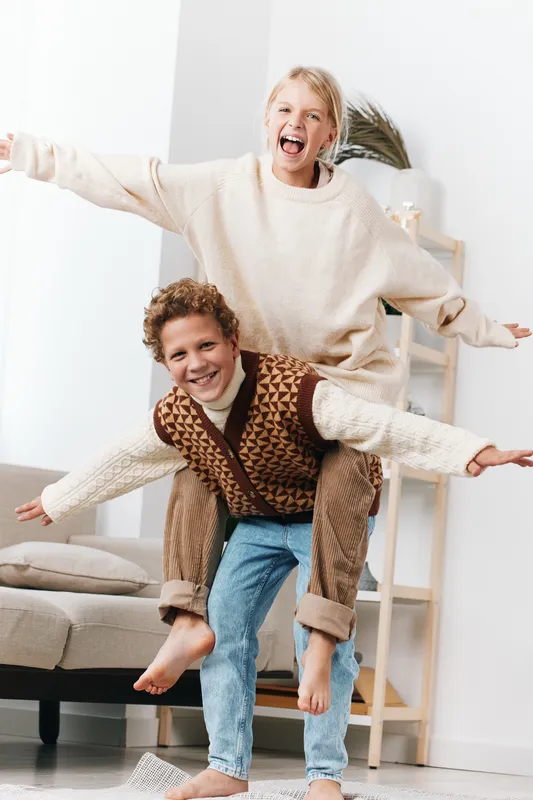 Two joyful children sit on an armchair, stacked with arms wide, sharing a moment of energetic laughter and carefree movement in a bright, cozy living space.