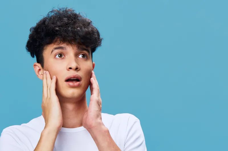 Young man with surprised expression wearing white t-shirt on blue background, showing shock and amazement with open mouth and wide eyes.