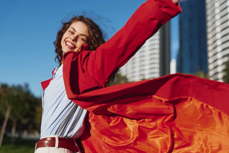Young woman with curly hair smiles as a red cape billows behind her in a sunlit outdoor park beside modern city buildings, her blue shirt and white jeans fluttering with the breeze, conveying freedom