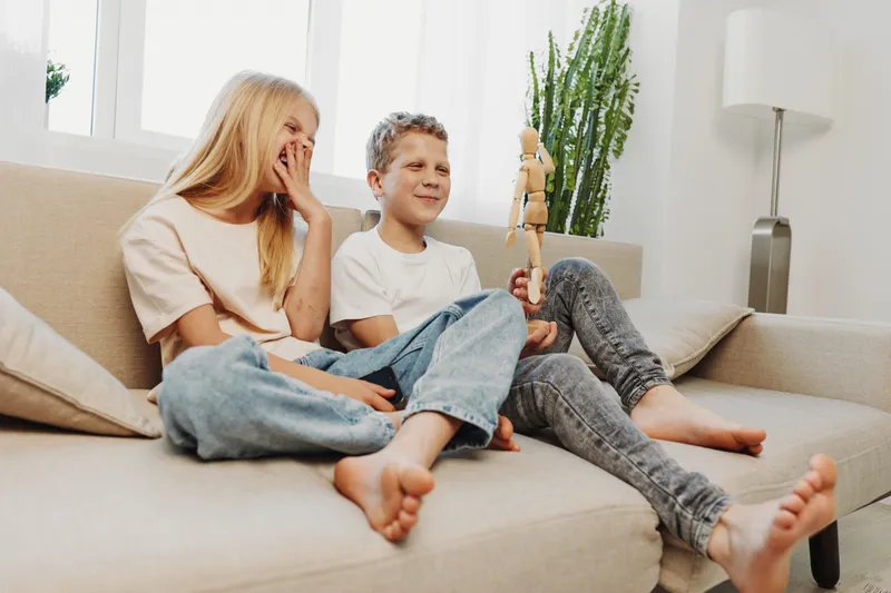 Happy children sitting barefoot on a sofa playing with a wooden toy, smiling and relaxed in a bright modern living room. Concept of childhood, family fun, and casual home life.