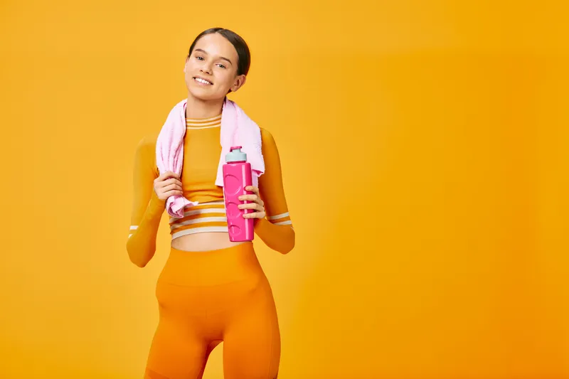 Woman in bright orange athletic wear with towel draped over shoulders and pink water bottle, ready for a workout against a bold orange backdrop, energetic and athletic.