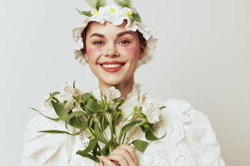 Cheerful young woman in a vintage floral bonnet holding fresh flowers, radiating joy and warmth in a light colored studio setting