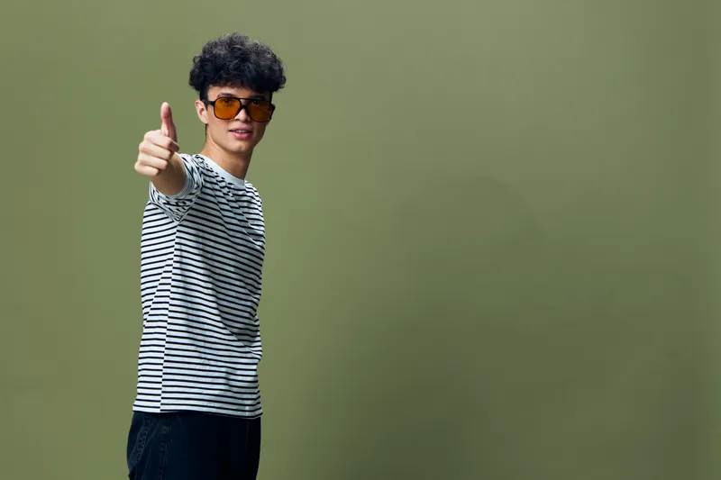 young man in striped shirt giving thumbs up gesture with confident smile on olive background, wearing sunglasses and casual black pants