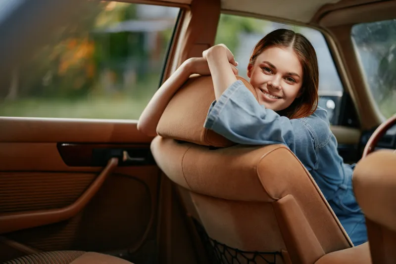 Cheerful young woman in a denim jacket smiling while sitting in a vintage car, summery atmosphere and warm lighting create a nostalgic vibe.
