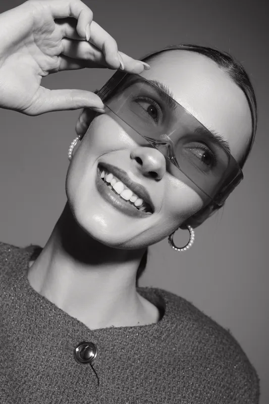 Smiling Caucasian woman with light skin wearing large sunglasses and hoop earrings, holding glasses frame in studio. Concept of fashion, style, and happiness.