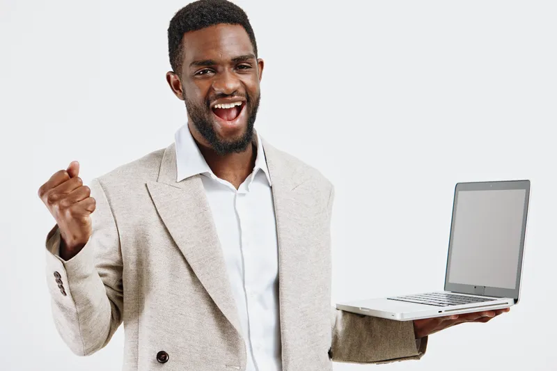Confident happy man of African descent holding a laptop, celebrating success, dressed in casual beige blazer and white shirt, isolated on plain white background, studio shot, positive energy concept