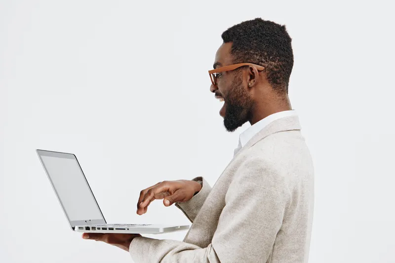 Close-up side profile of a smiling man with dark skin, wearing glasses and a beige sweater, using a laptop on a plain white background, representing technology, professional work, and online communica