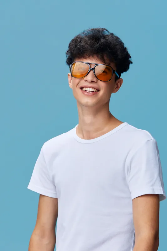 smiling young man with curly hair wearing two pairs of sunglasses and a white t-shirt against a blue background expressing fun and carefree mood