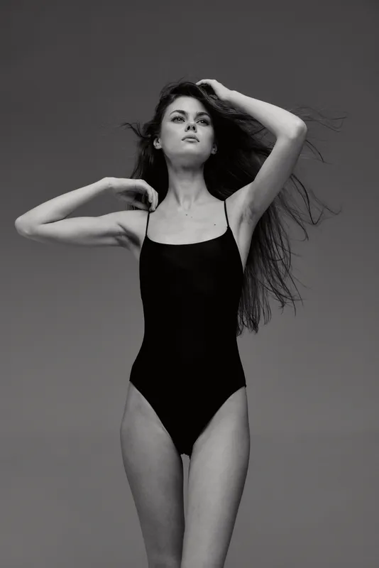 Black and white portrait of a slender woman posing in a swimsuit with flowing hair and confident expression against a plain background.