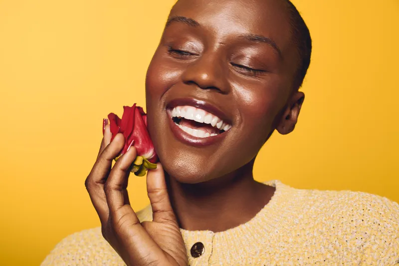 Smiling dark skinned African woman with closed eyes holds red flower near face on yellow background wearing knitted beige sweater. Concept for beauty, happiness, nature, and self-care.