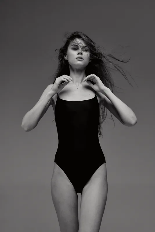 Black and white portrait of elegant woman in black swimsuit with windblown hair and confident expression against plain background.
