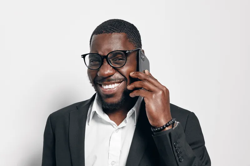 African American man with dark skin wearing glasses and black suit talking on smartphone smiling in studio. Portrait of confident businessman with beard and white shirt on white background.