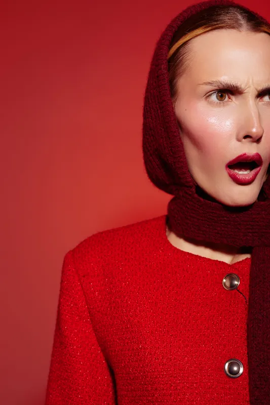 Surprised woman in red coat and matching scarf looks shocked against red background. Close-up portrait with expressive face and dramatic lighting. Concept of emotion and fashion.