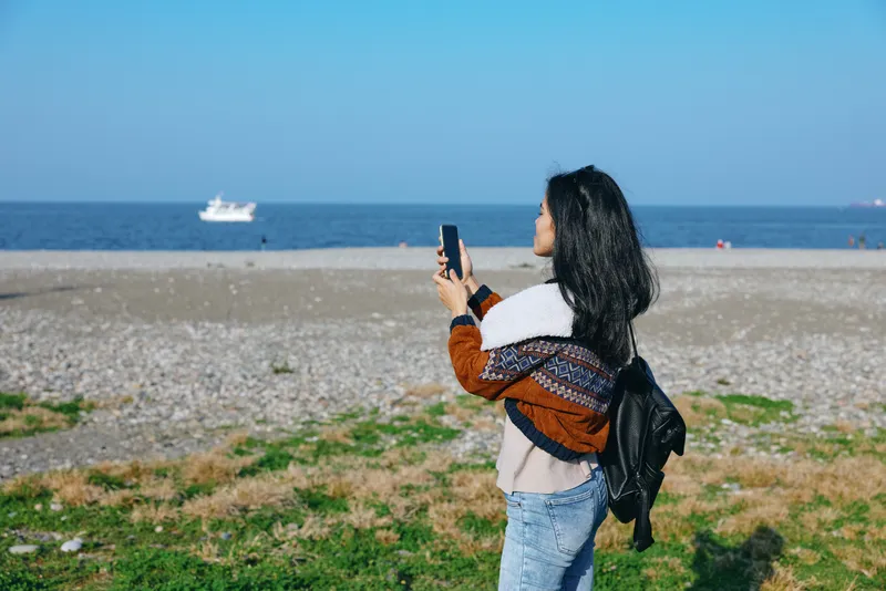 Woman on a beach holds smartphone, capturing sea and horizon in bright daylight, with backpack and casual attire, a relaxed outdoor photography scene by the coast.