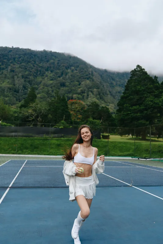 Young athletic woman running on a tennis court in a white outfit, joyful expression, surrounded by greenery and mountains, perfect for fitness and sports themes