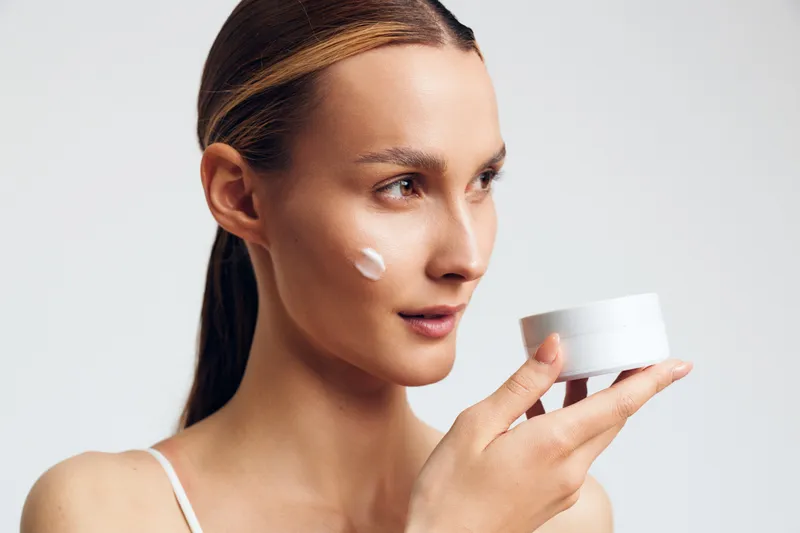 A young woman with clear skin holds a white cosmetic jar and applies cream to her cheek, demonstrating skincare routine and facial care in a bright studio setting.