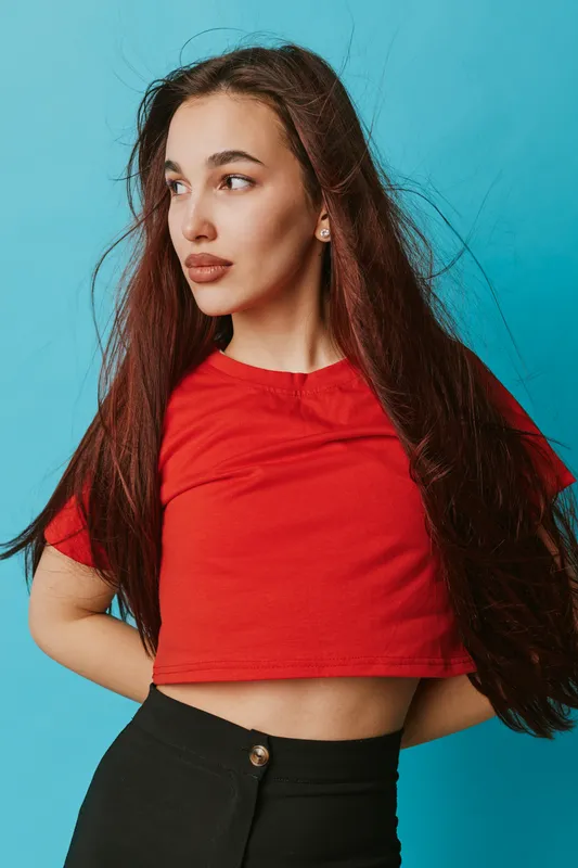 Young woman with long brown hair wearing red crop top and black pants posing and looking sideways on blue background. Casual fashion, beauty, confident female model with natural makeup and earrings.
