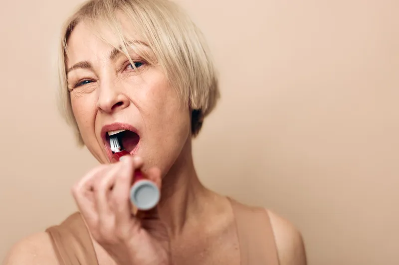 Mature woman with short blonde hair applies lipstick in front of beige background. Close-up portrait showing natural makeup routine and beauty care concept for older adults.