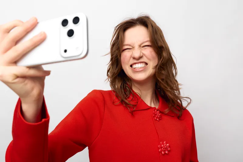 Young woman in red sweater takes a selfie with smartphone, smiling and squinting eyes. White background, casual style, concept of social media, communication, and modern technology.