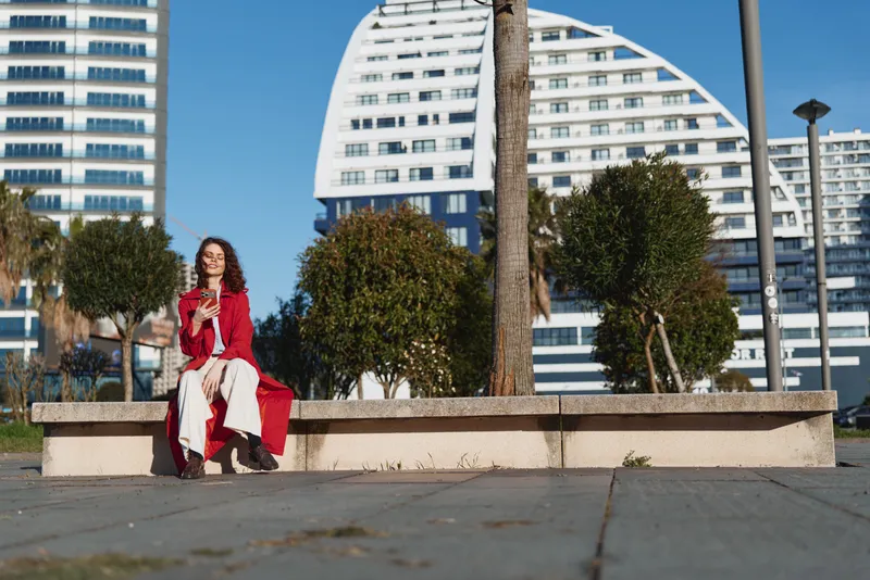 Cheerful woman outdoors, red coat, sitting on a bench in an urban plaza, sunny day, relaxed pose, modern buildings in background, natural colors