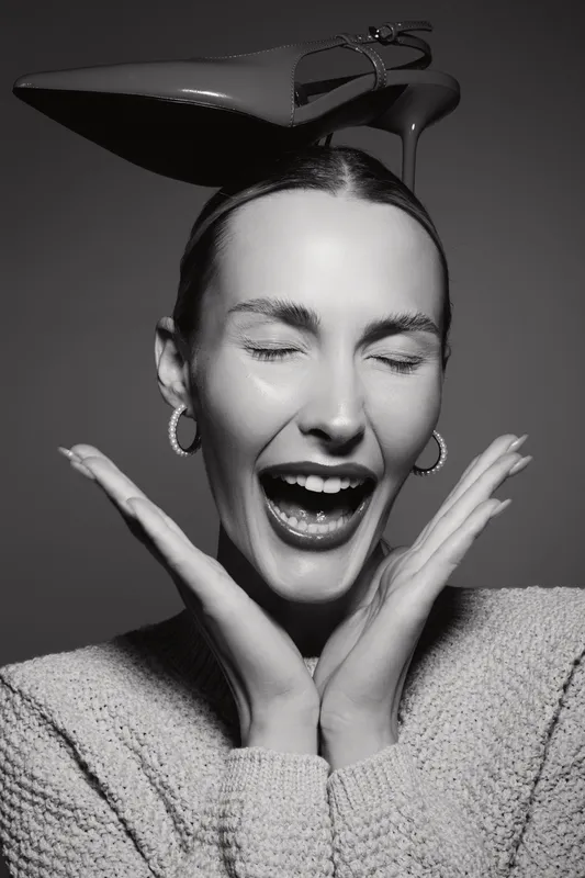 Happy Caucasian woman with fair skin and closed eyes laughs with shoe balanced on head, wearing sweater and hoop earrings. Concept of joy and playful fashion.