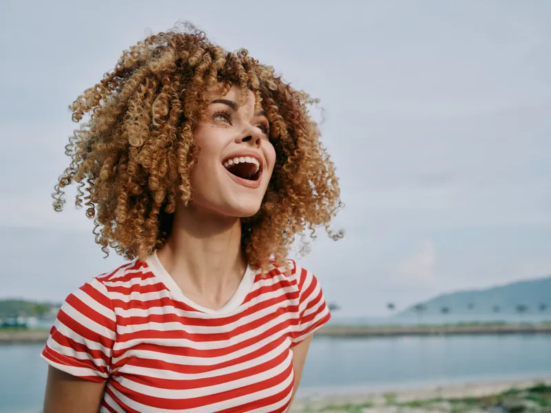 Joyful young woman with curly hair wearing a red striped shirt stands outdoors by a blue lake, smiling broadly as sunlight highlights her radiant expression.