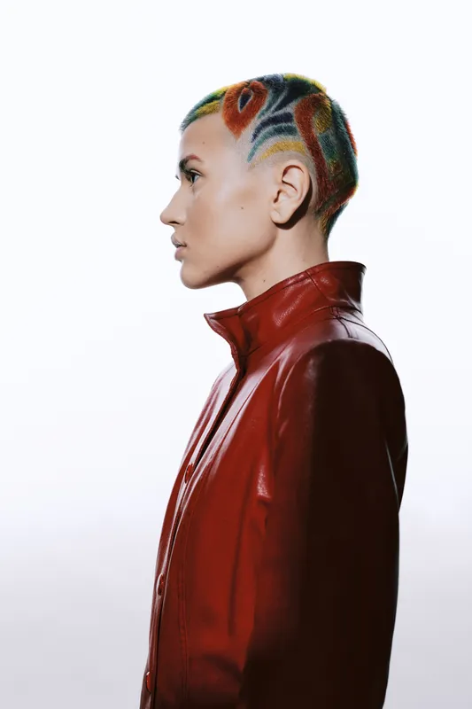 Profile of a young woman with colorful shaved hair styled in artistic patterns, wearing a red leather jacket against a bright white background, showcasing bold fashion and creativity.