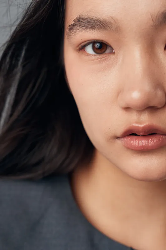 Young woman closeup portrait with natural skin makeup and black hair, looking at camera with neutral expression in studio lighting, detailed face and lips visible.