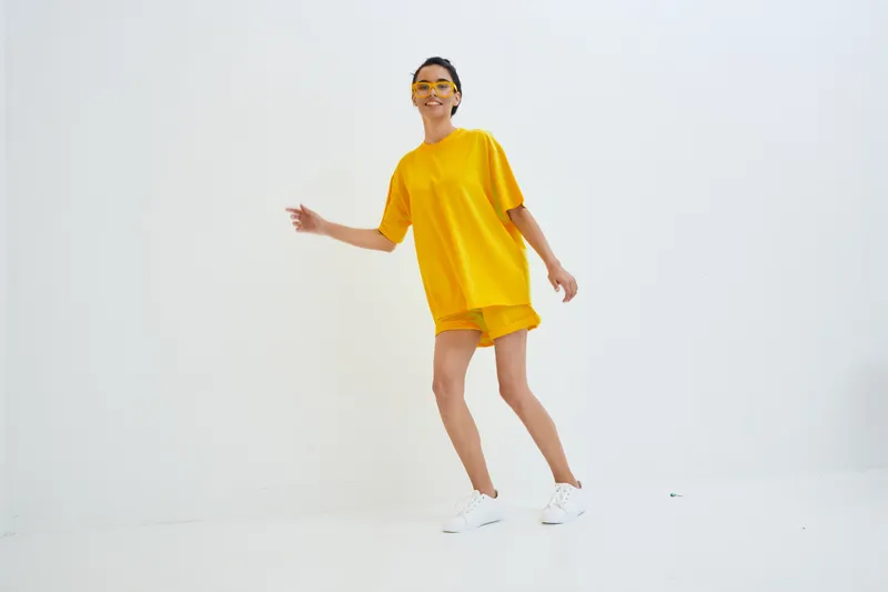 young woman in bright yellow outfit smiling and posing against white background, wearing casual shorts and sunglasses indoors, joyful and energetic mood
