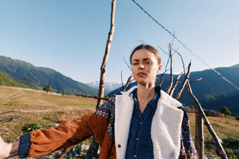 Woman portrait in mountains and rural landscape near a wooden fence wearing layered jacket and coat, standing in meadow under blue sky with calm expression and autumn sunlight.