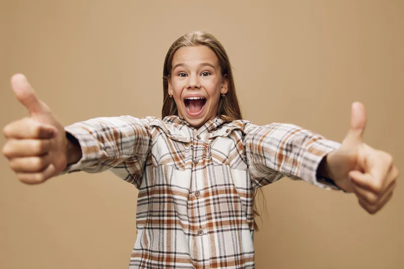 Girl with long hair and plaid shirt showing thumbs up with excited facial expression and open mouth on beige background for positivity and approval.